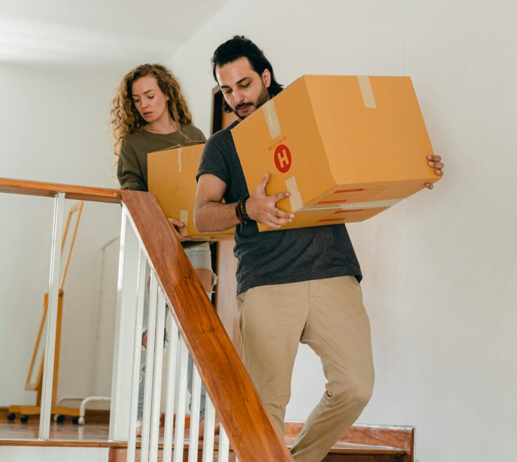 Full body of young woman and barefoot ethnic man in casual clothes carrying boxes on wooden stairs while moving into new home